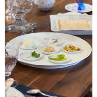A Louvre Seder Dishes Set of 6 by Bernardaud displays Passover foods—egg, parsley, romaine, charoset, horseradish, and shank bone—on a wooden table set for a meal with matzah in the background.