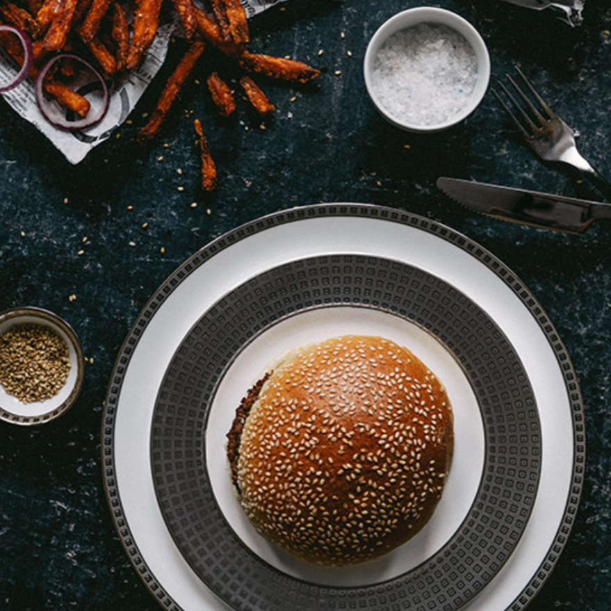 A sesame seed burger bun rests on a Bernardaud Athena Platinum Dinnerware plate, surrounded by bowls of salt and sesame seeds, carrot fries, and utensils on a dark table.