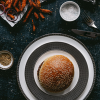 A sesame seed burger bun rests on a Bernardaud Athena Platinum Dinnerware plate, surrounded by bowls of salt and sesame seeds, carrot fries, and utensils on a dark table.