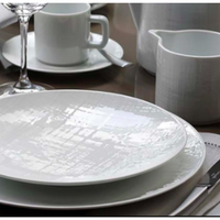 A close-up of a table set with Bernardaud's Organza White Dinnerware—plates, cup and saucer, wine glass, jug, and teapot—showcasing subtle textured patterns that echo Limoges porcelain elegance in a minimalist setting.