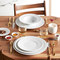 A wooden dining table set with Bernardaud Twist White Dinnerware, gold cutlery, clear wine glasses, small bowls of spices and nuts, and a lit white candle in a decorative holder. Two wooden chairs are visible.