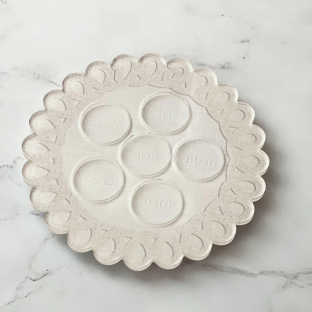 The Lucite Scalloped Seder Plate by BT- Shalom, featuring a round white design with scalloped edges and six Hebrew-labeled circles, creates a modern centerpiece for your Passover celebration when displayed on a marble surface.