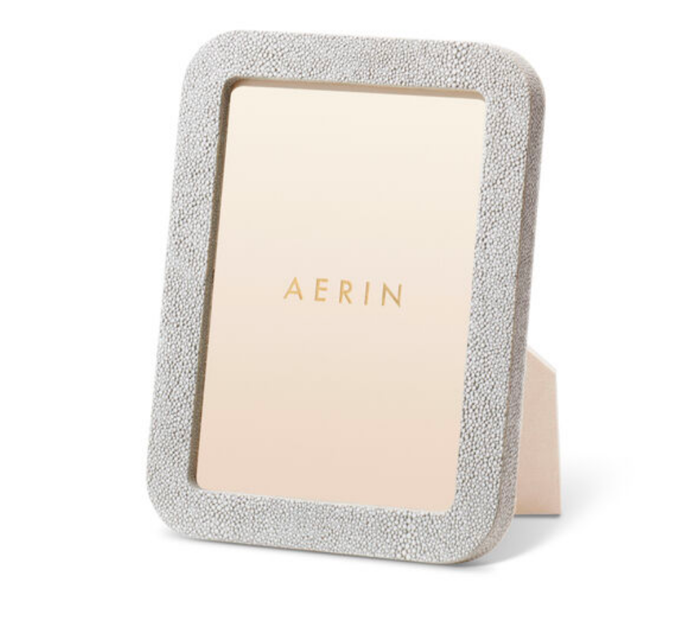 The Modern Shagreen Frame Dove by Aerin is a rectangular, textured light gray embossed shagreen frame with a stand, featuring a placeholder displaying AERIN in gold letters on a beige background.
