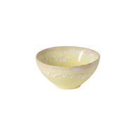 The Costa Nova Taoromina Dinnerware Yellow is a round ceramic bowl featuring a pale yellow base with a wavy pastel pink and cream glaze near the rim, highlighting artisanal craftsmanship, set against a white background.