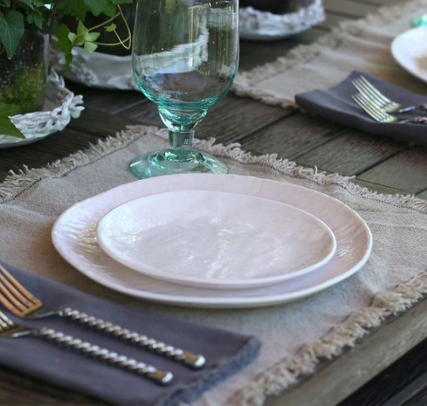 A close-up of a rustic table set with Relish's Weave Melamine Dinnerware in white, displayed on a beige placemat atop a wooden surface, with cutlery on a gray napkin, clear water glass, and plants in the background.
