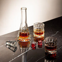 A Saint-Louis Manhattan Barware glass decanter and two whiskey glasses rest on dark marble with red dice and crystal stoppers, illuminated in soft light.