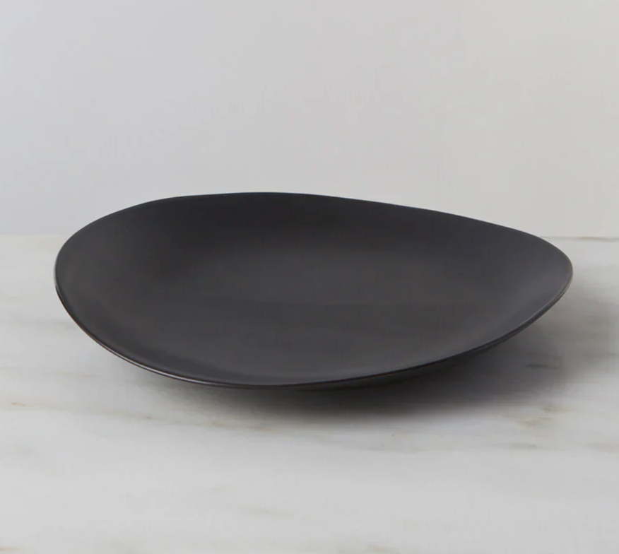 The Simon Pearce Barre Dinnerware Slate plate, featuring a matte black, asymmetrical stoneware design, rests on light marble with a plain white background—highlighting the refined style of modern dinnerware.