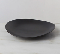 The Simon Pearce Barre Dinnerware Slate plate, featuring a matte black, asymmetrical stoneware design, rests on light marble with a plain white background—highlighting the refined style of modern dinnerware.