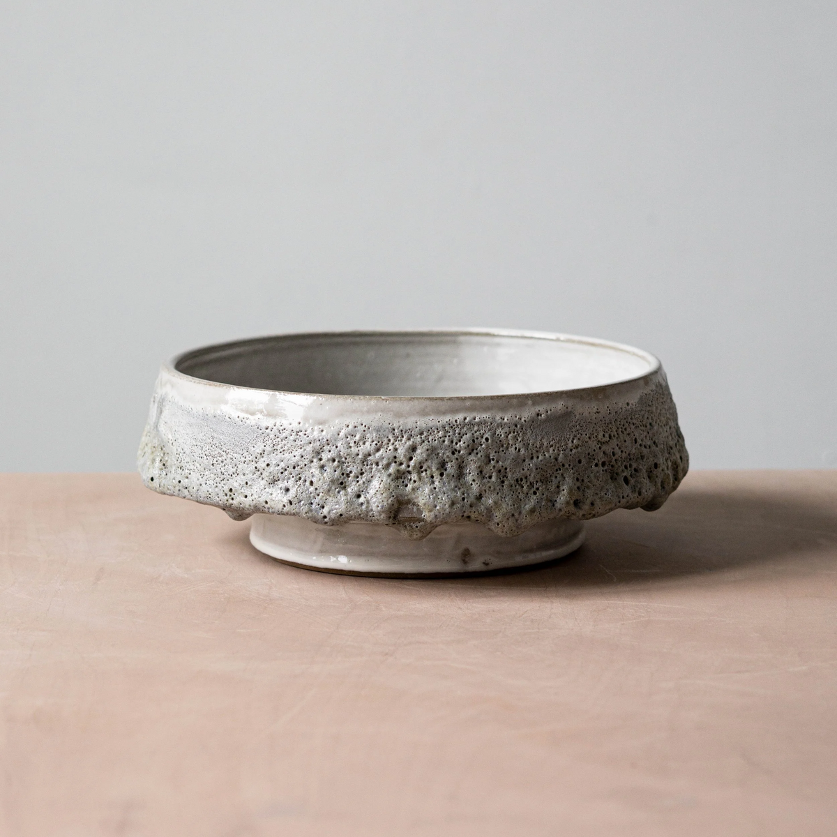 The Cindy Froth Vase by Kalya OD Studio features a wide, shallow ceramic shape with a textured, uneven rim and speckled glaze. Its rustic, handmade look makes it a perfect addition to artisan home decor. Displayed on a light pink surface against gray.
