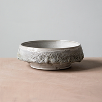 The Cindy Froth Vase by Kalya OD Studio features a wide, shallow ceramic shape with a textured, uneven rim and speckled glaze. Its rustic, handmade look makes it a perfect addition to artisan home decor. Displayed on a light pink surface against gray.