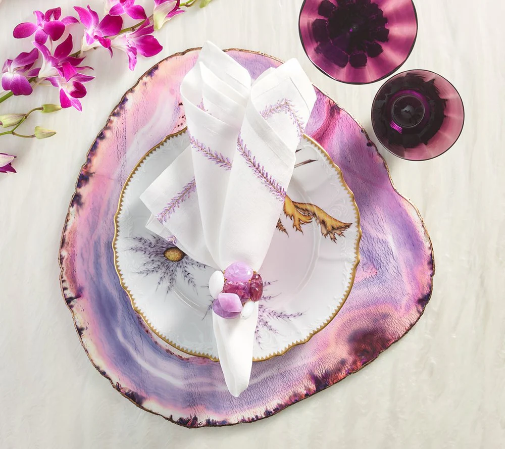 A table setting with a white and purple plate on a shell-shaped charger, white napkin with lavender embroidery, Kim Seybert Sea Stone Napkin Ring in Lilac, two purple glasses, and purple orchids on a white surface.