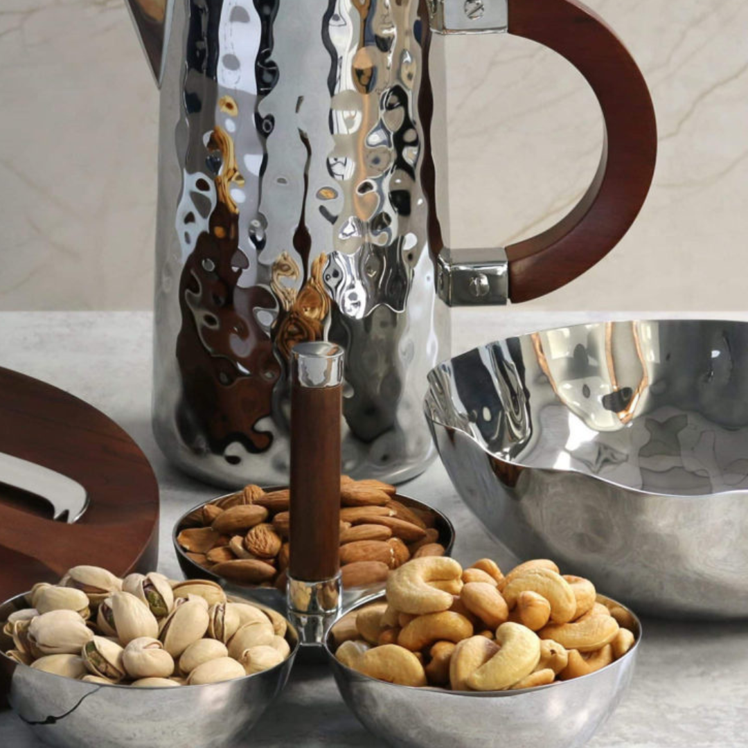 A set of hammered metal serveware with wooden handles holds almonds, cashews, and pistachios. A matching Mary Jurek Blossom Stainless Steel Bowl sits on a light marble surface, reflecting the snacks and surroundings.