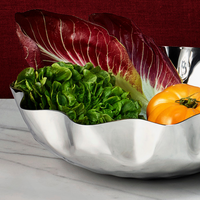 A Mary Jurek Blossom Stainless Steel Bowl sits on marble, filled with green lettuce, red radicchio, and two yellow tomatoes against a deep red backdrop.