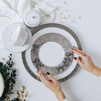 With manicured black nails, someone arranges Bernardaud Silva Dinnerware Serving Pieces—elegant white and silver plates with platinum relief—on a table set with a teacup, saucer, spoon, greenery, and gold leaf confetti.