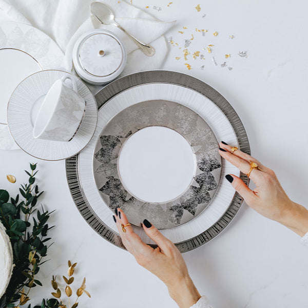 With manicured black nails, someone arranges Bernardaud Silva Dinnerware Serving Pieces—elegant white and silver plates with platinum relief—on a table set with a teacup, saucer, spoon, greenery, and gold leaf confetti.