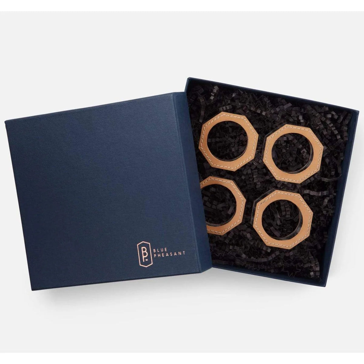 A navy blue gift box with Blue Pheasant branding opens to reveal four Arthur Leather Camel Napkin Rings, stylishly arranged on black shredded paper—an elegant addition to any table setting.