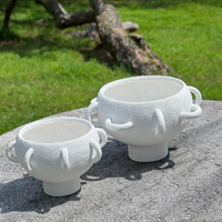 Two BT-Shalom Textured Vessel White porcelain pieces with a unique multi-handle design are displayed outdoors on stone, set against green grass and a blurred fallen tree in the background.