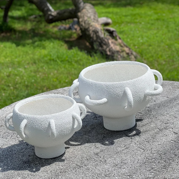 Two BT-Shalom Textured Vessel White porcelain pieces with a unique multi-handle design are displayed outdoors on stone, set against green grass and a blurred fallen tree in the background.