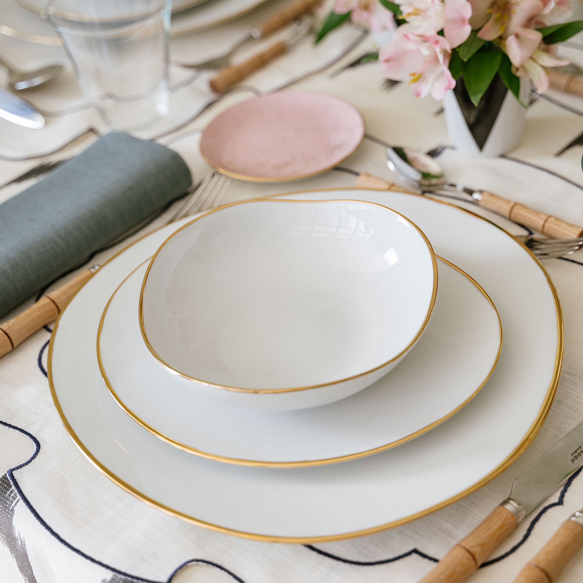 A close-up of an elegant table set with Molecot's Simply Gold Dinnerware—white bowl, salad plate, and dinner plate with gold trim—paired with bamboo-handled cutlery, a green napkin, and pink flowers in a vase.