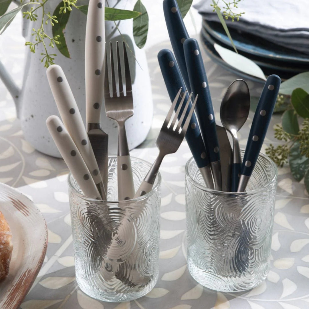 Two glass tumblers hold Blue Pheasant's Jones Flatware Ivory—a modern cutlery set with ivory handles—displayed alongside a jug, plates, and leafy greenery on a patterned table. All utensils are dishwasher safe.