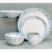 The Costa Nova Brisa Blue Dinnerware set features two plates, a bowl, and a mug in white with blue and tan watercolor edges, displayed on a gray surface. This stoneware set is both elegant and versatile.