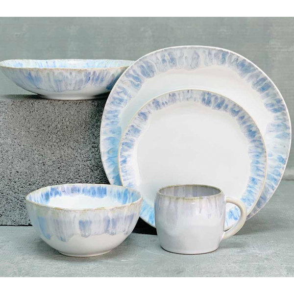 The Costa Nova Brisa Blue Dinnerware set features two plates, a bowl, and a mug in white with blue and tan watercolor edges, displayed on a gray surface. This stoneware set is both elegant and versatile.