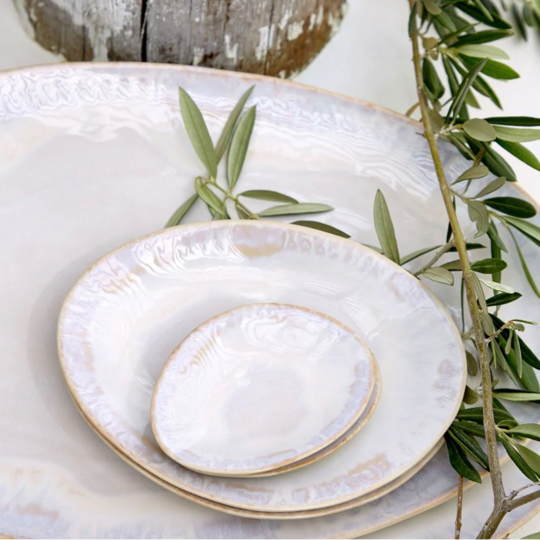Three stacked Brisa Sal Dinnerware plates by Casafina/Costa Nova, with a soft iridescent glaze, are topped with green olive branches on a large platter and set on a rustic wooden surface inspired by the Algarve coast.