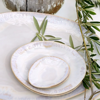 Three stacked Brisa Sal Dinnerware plates by Casafina/Costa Nova, with a soft iridescent glaze, are topped with green olive branches on a large platter and set on a rustic wooden surface inspired by the Algarve coast.