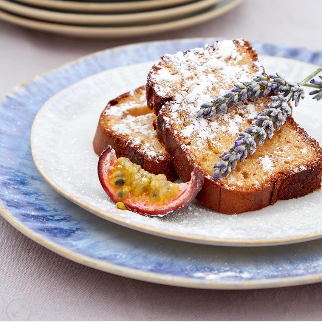 Two slices of pound cake with powdered sugar, lavender, and half a passion fruit, served on a white plate with blue rim from the Brisa Blue Dinnerware by Costa Nova. Made in Portugal.