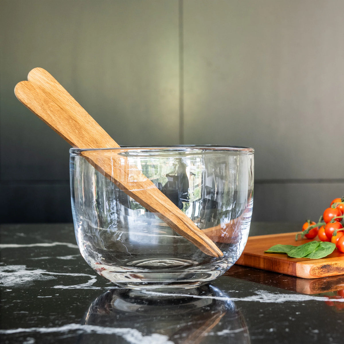 The Burlington Bowl by Simon Pearce, handmade from glass, features two wooden salad servers and sits on a black marble countertop. Nearby, a wooden board with cherry tomatoes and spinach highlights its organic design for your kitchen.