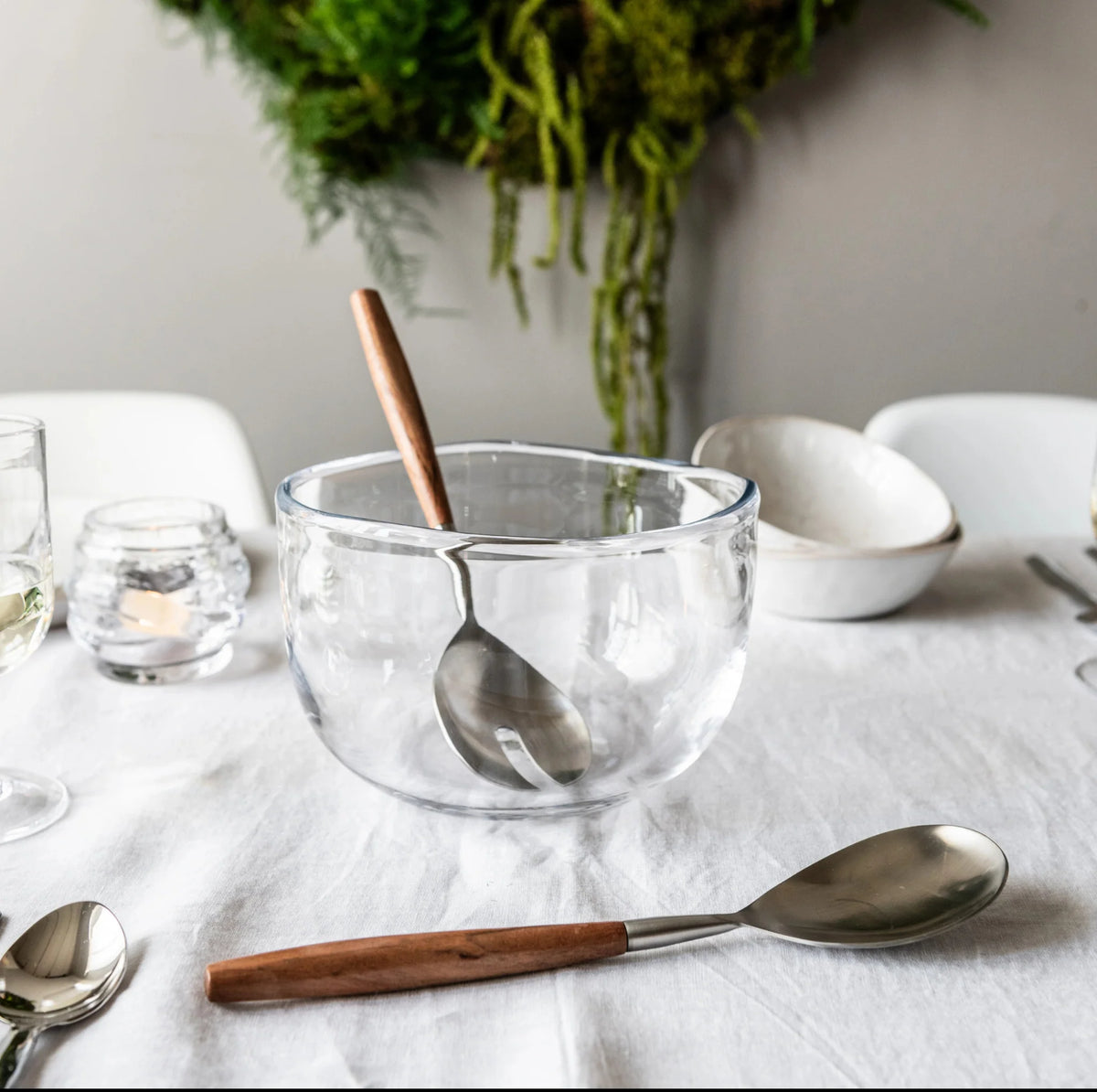 The Burlington Bowl by Simon Pearce, paired with a wooden-handled serving spoon, rests on a white tablecloth amid cutlery, a candle holder, and bowls, while greenery hangs in the background.