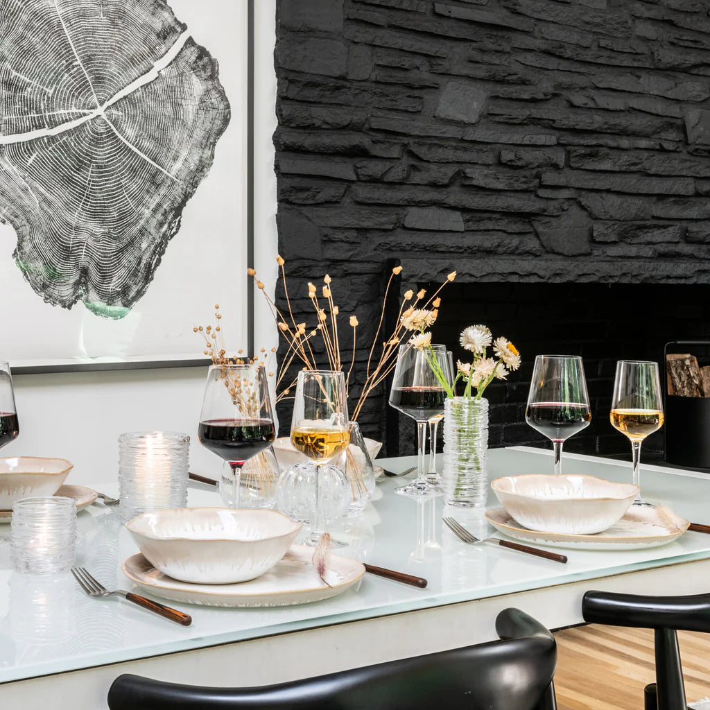 A modern dining table set with Simon Pearce Burlington Dinnerware Bluff—featuring glazed edge plates and bowls—complemented by wine glasses, candles, and simple floral arrangements, set against a black stone fireplace and a large framed tree-ring artwork.