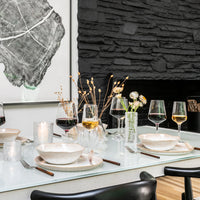 A modern dining table set with Simon Pearce Burlington Dinnerware Bluff—featuring glazed edge plates and bowls—complemented by wine glasses, candles, and simple floral arrangements, set against a black stone fireplace and a large framed tree-ring artwork.