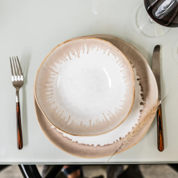 A table setting featuring a fork, knife, and spoon, a beige-rimmed Burlington Dinnerware Bluff bowl with a beautifully glazed edge on a matching plate by Simon Pearce, alongside a glass of red wine on a white surface.