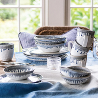 Simon Pearce Burlington Dinnerware Pool—a collection of ceramic dishes, bowls, and mugs with blue and gray glaze—rests on blue and white cloths atop a table near a window overlooking greenery.