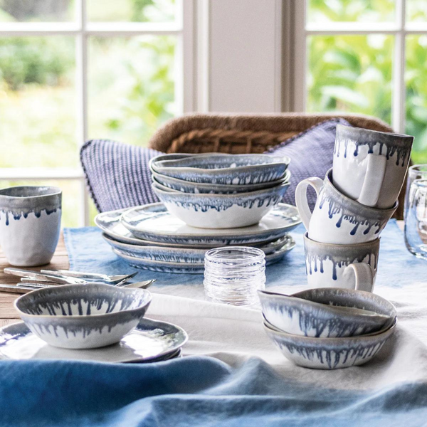 Simon Pearce Burlington Dinnerware Pool—a collection of ceramic dishes, bowls, and mugs with blue and gray glaze—rests on blue and white cloths atop a table near a window overlooking greenery.