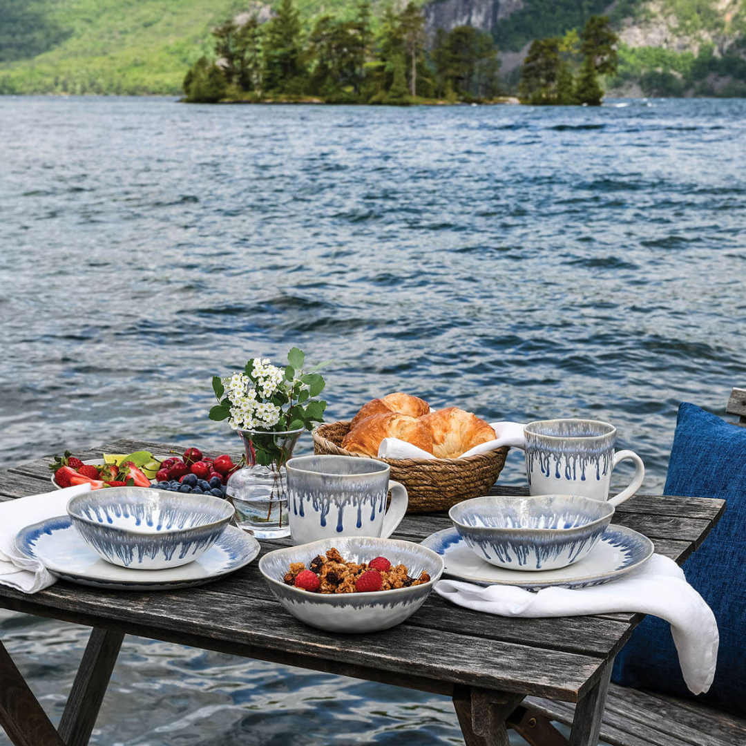 A rustic wooden table by a lake with mountain views is set with Simon Pearce Burlington Dinnerware Pool, featuring a glazed edge, alongside croissants, fruit, and flowers.