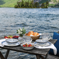 A rustic wooden table by a lake with mountain views is set with Simon Pearce Burlington Dinnerware Pool, featuring a glazed edge, alongside croissants, fruit, and flowers.