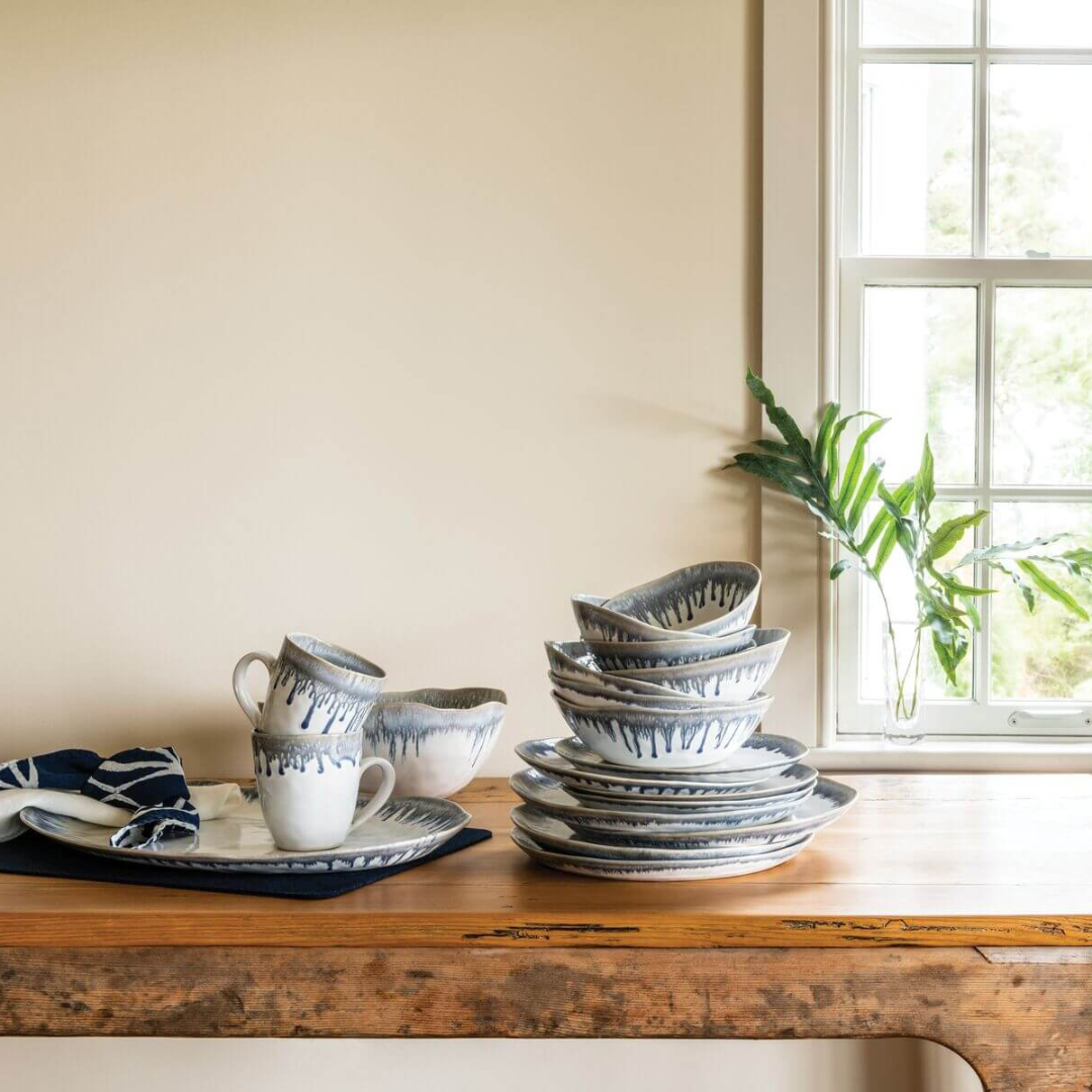 Simon Pearce’s Burlington Dinnerware Pool set—white ceramic dishes with a blue glazed edge—is arranged on a wooden table by a window. The unique set includes plates, bowls, and mugs.