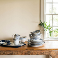 Simon Pearce’s Burlington Dinnerware Pool set—white ceramic dishes with a blue glazed edge—is arranged on a wooden table by a window. The unique set includes plates, bowls, and mugs.