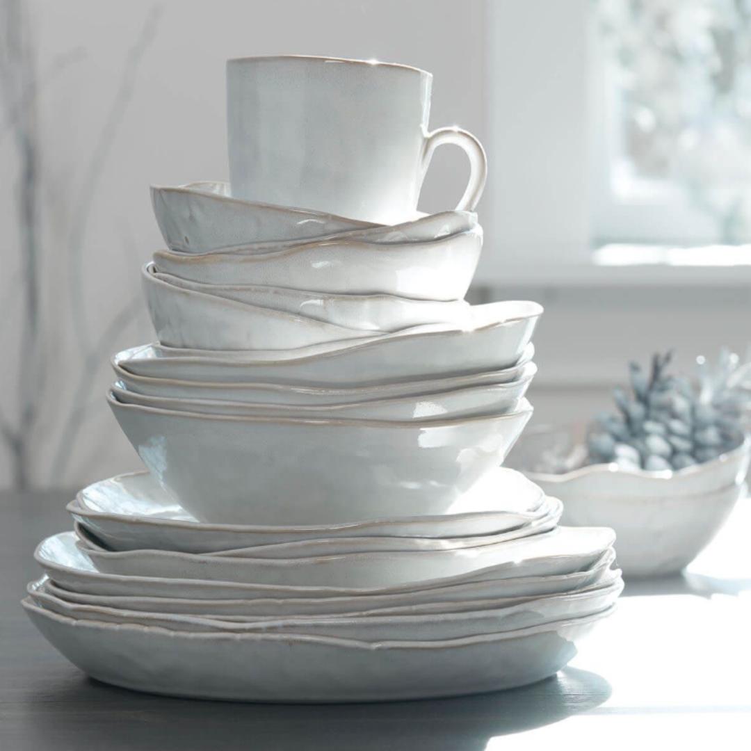 A stack of Simon Pearce Burlington Dinnerware Cloud—handmade white ceramic plates and bowls with organic shapes—rests on a table in bright light, with a blurry bowl of succulents by a sunny window.