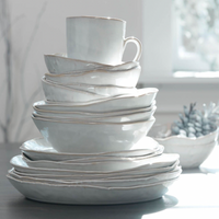 A stack of Simon Pearce Burlington Dinnerware Cloud—handmade white ceramic plates and bowls with organic shapes—rests on a table in bright light, with a blurry bowl of succulents by a sunny window.