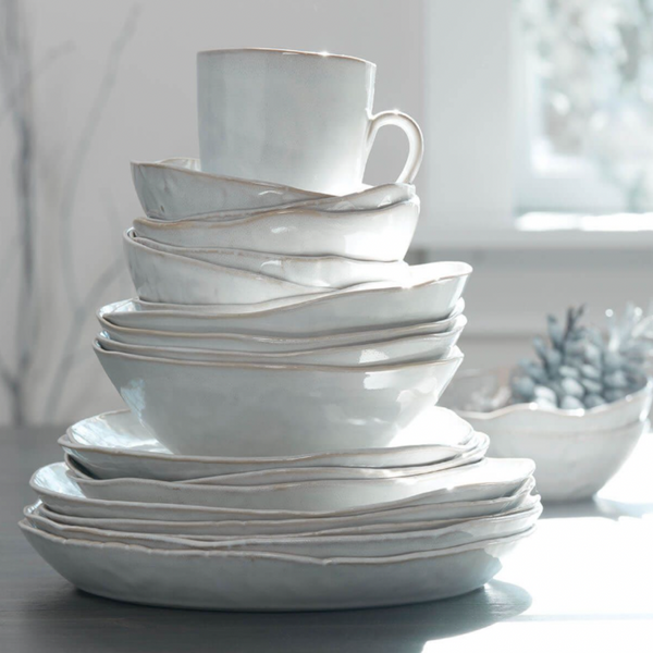 A stack of Simon Pearce Burlington Dinnerware Cloud—handmade white ceramic plates and bowls with organic shapes—rests on a table in bright light, with a blurry bowl of succulents by a sunny window.