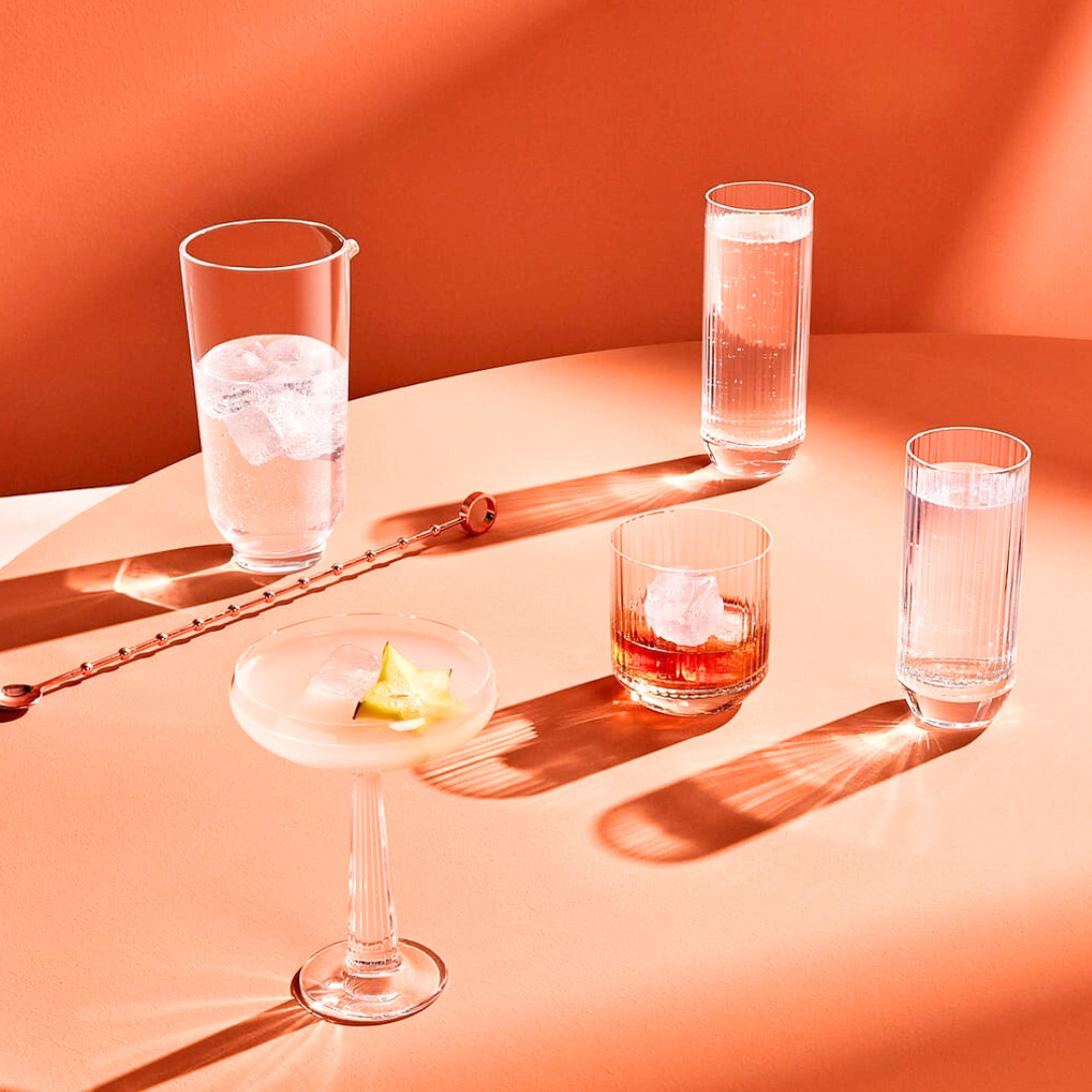 Four cocktails in sleek, minimalist Nude Glassware from the Big Top Barware Set sit on a peach table, each with ice and garnishes, casting bold shadows in a bright modern setting.