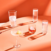 Four cocktails in sleek, minimalist Nude Glassware from the Big Top Barware Set sit on a peach table, each with ice and garnishes, casting bold shadows in a bright modern setting.