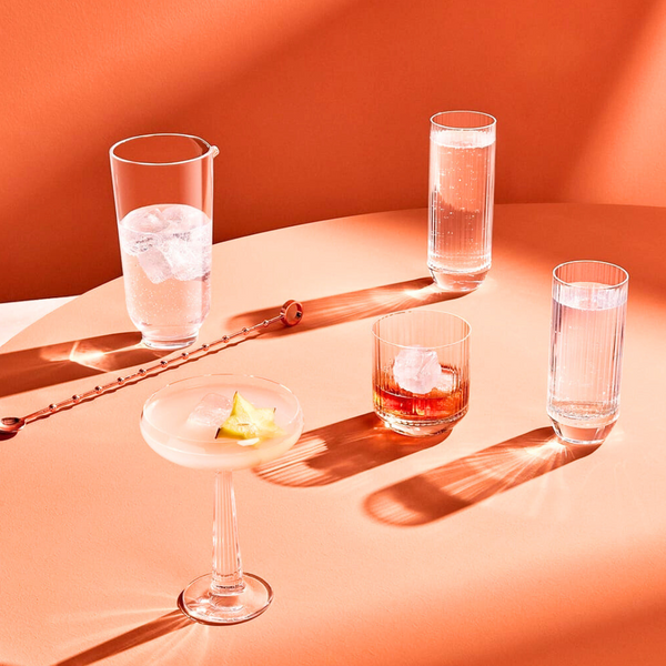 Four cocktails in sleek, minimalist Nude Glassware from the Big Top Barware Set sit on a peach table, each with ice and garnishes, casting bold shadows in a bright modern setting.