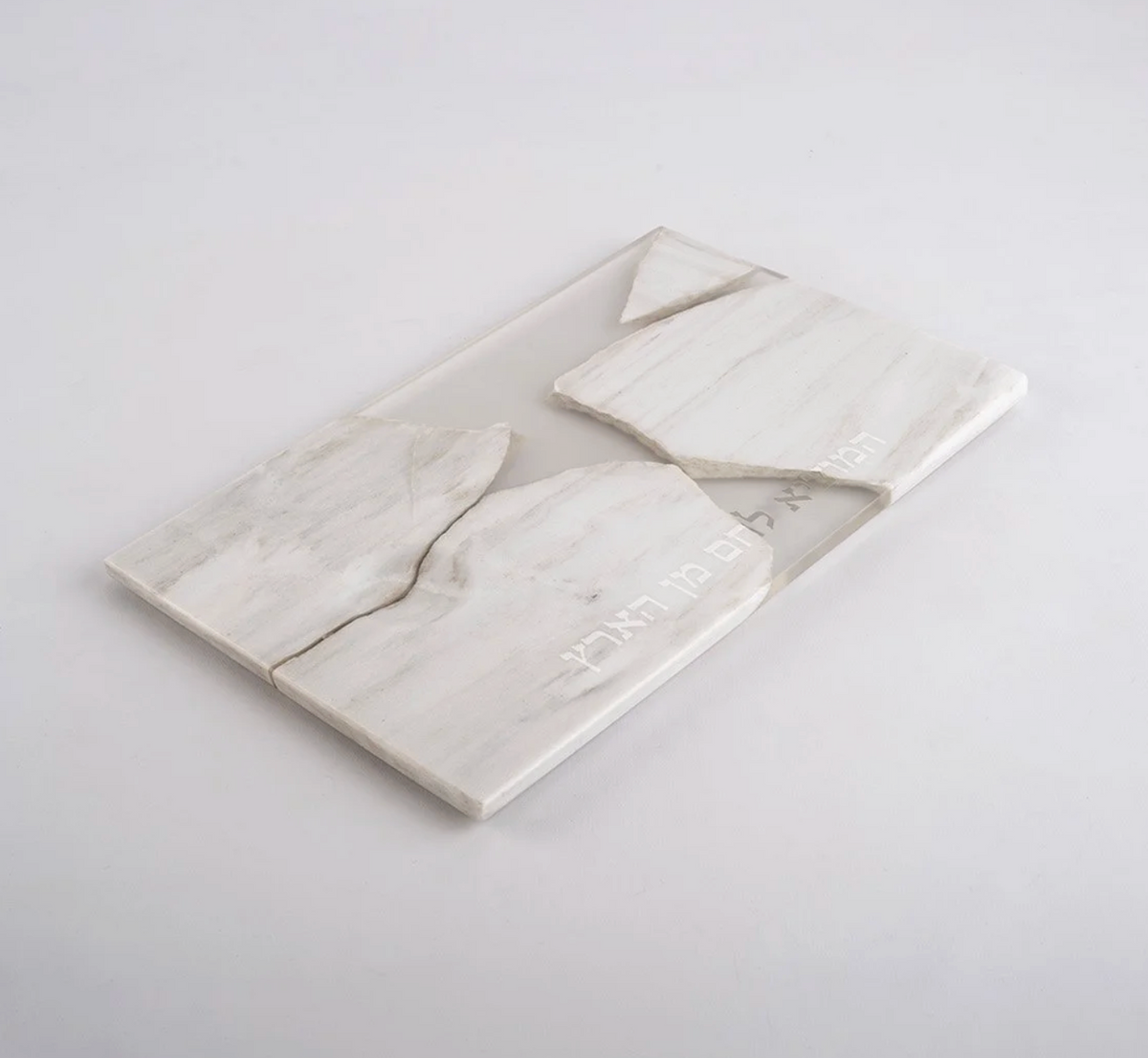 The Mutual Challah Board w/ Hebrew Blessing by CDMX / BRUCI features white marble pieces fitted together, with partially visible Hebrew text engraved along the cracks, evoking Shabbat gatherings.
