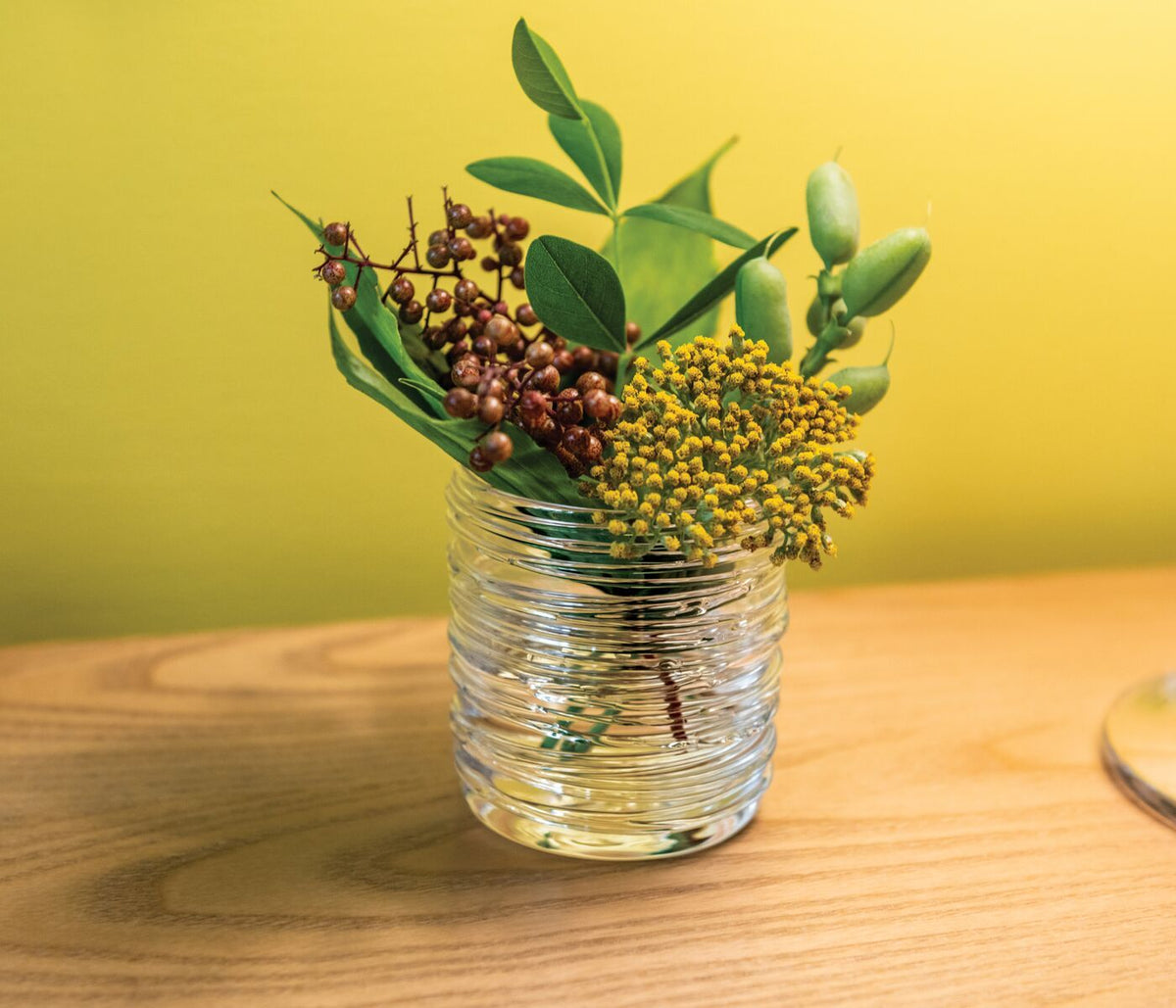 The Echo Lake Tealight In Gift Box by Simon Pearce features clear, textured glass and is displayed on a wooden surface with green leaves, yellow flowers, and red berries set against a yellow background.
