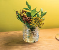 The Echo Lake Tealight In Gift Box by Simon Pearce features clear, textured glass and is displayed on a wooden surface with green leaves, yellow flowers, and red berries set against a yellow background.