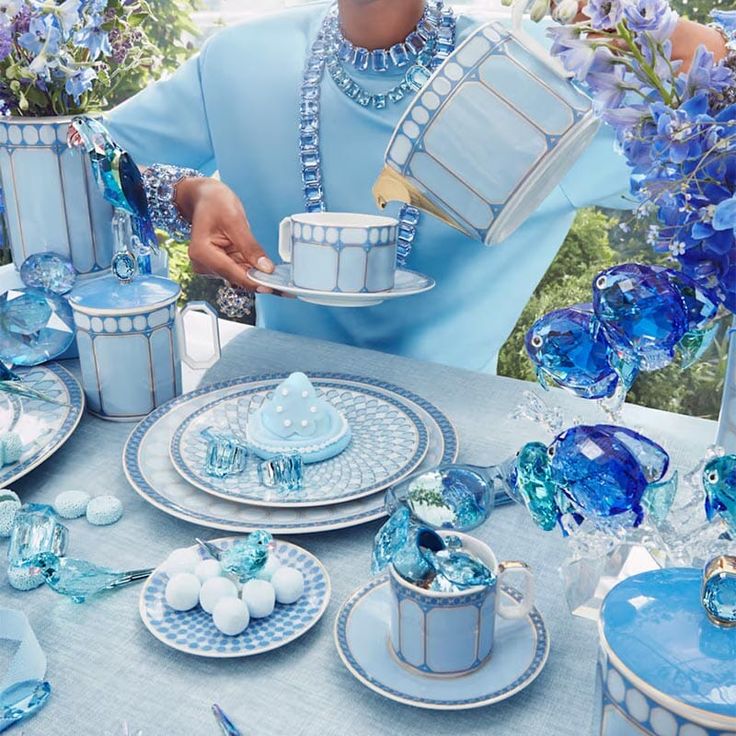 A person in blue pours cream into a Swarovski Signum Azure Blue Mug by Swarovski x Rosenthal at an elegantly set table of coordinated blue-themed décor, featuring sparkling glass ornaments, flowers, and pastel sweets for a luxurious tea party.